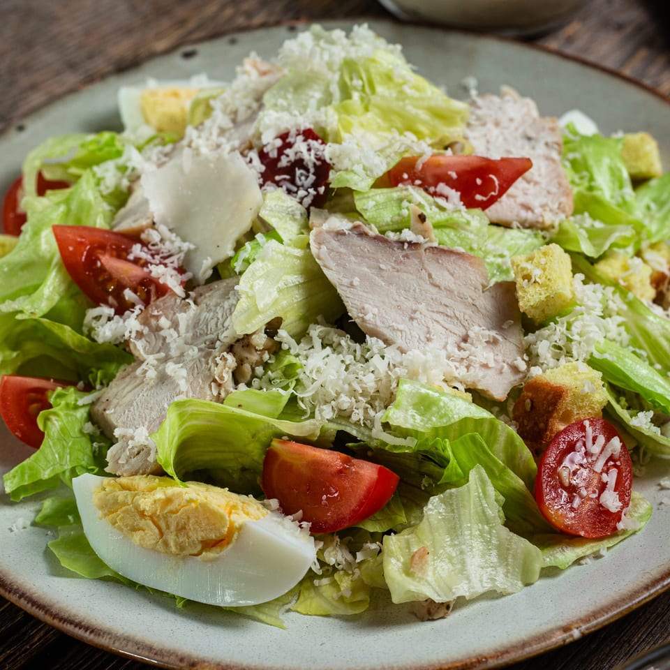 Close-up of a Caesar salad with chicken, tomatoes, hard-boiled egg, croutons, and shredded cheese.